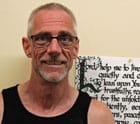 The author of the story, Patrick, looks at the camera and smiles. He is a man in his mid-fifties with glasses, short grey hair, and a small beard.Behind him is a hand-lettered sign with a prayer from St. Francis.